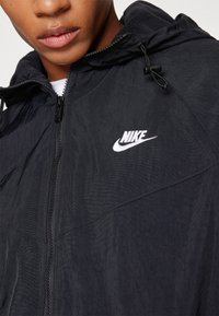 Gros plan d'une personne portant une veste à capuche noire Nike avec un logo blanc sur la poitrine et une fermeture éclair partiellement fermée.