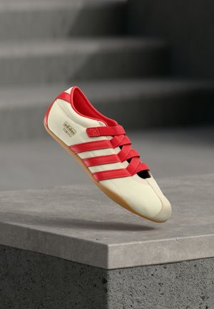 Crvena i krem sportska cipela s tri crvene pruge, mekanim gornjim dijelom, gumenim potplatom i remenčićima na čičak. Ima elegantan dizajn niskog profila.