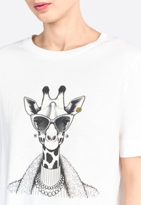 T-shirt bianca con una grafica in bianco e nero di una giraffa che indossa occhiali da sole, perle e un colletto peloso, con dettagli e accenti intricati.