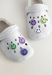 Weiße Clogs mit weichem Innenfutter, versehen mit vier dekorativen Smiley-Anhängern in Grün, Lila, Blau und mit gemusterten Akzenten.