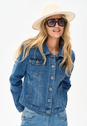Blondynka w białym kapeluszu z szerokim rondem, ogromnych ciemnych okularach przeciwsłonecznych, niebieskiej kurtce dżinsowej i jasnych dżinsach, lekko się uśmiecha.