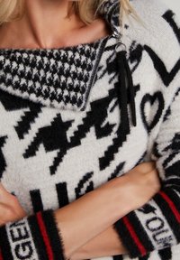 Pull à motifs en noir et blanc avec un col à fermeture éclair, une texture douce, et des manches larges rayées comportant des accents rouges et des détails textuels.