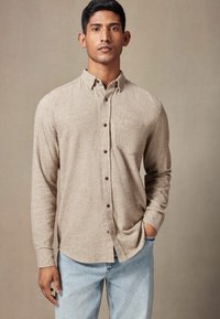 Beige skjorte med knapper og tekstureret stof, enkelt brystlomme og brune knapper. Sat sammen med lyseblå jeans, poseret foran en neutral baggrund.