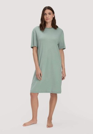 Frau steht barfuß in einem lockeren, knielangen, hellgrünen Kurzarmkleid vor einem schlichten Hintergrund.