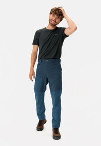 Heren buitenskleding met een donkergrijs T-shirt en blauwe broek met meerdere zakken, ritsdetails en een comfortabele pasvorm. Bruine wandelschoenen.