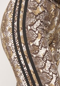 Bruna och beige leggings med ormskinsmönster; har tre svarta ränder längs sidan. Tillverkade av stretchig, texturerad tyg.