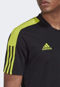 Detailní záběr na osobu v černém sportovním tričku Adidas s neonově žlutými ramenními panely, třemi pruhy na rukávu a logem Adidas na hrudi.