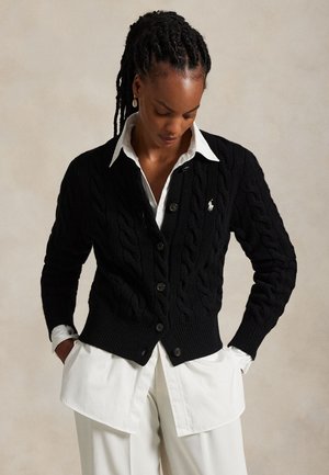 Mujer con el pelo trenzado que lleva un cárdigan negro de punto con botones sobre una camisa blanca y pantalones blancos, de pie con las manos en los bolsillos.