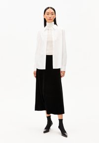 Weißes Hemd, cremefarbener Rollkragenpullover, schwarzer Samtmidirock und elegante schwarze Stiefeletten, vor einem schlichten weißen Hintergrund.