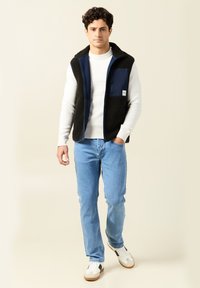 Fleeceweste in Schwarz und Marineblau mit Reißverschlusstasche, weißem Pullover, blauen Jeans und weißen Sneakers mit braunen und schwarzen Akzenten.