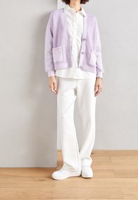 Lavendelstickad cardigan med ribbat mönster, två fickor fram, knäppning med knappar. Bärs över en vit skjorta och tillsammans med vita byxor och sneakers.