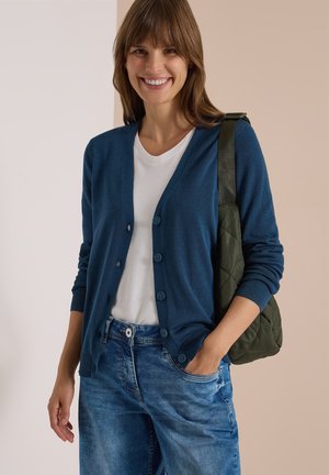 Femme souriante portant un cardigan bleu sur un t-shirt blanc et un jean, portant un sac à bandoulière matelassé vert, une main dans la poche.