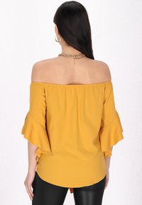 Mosterdkleurige off-the-shoulder top met ruches aan de mouwen, zachte stof en een losse pasvorm, gecombineerd met zwarte, strak aansluitende broek. Achteraanzicht weergegeven.