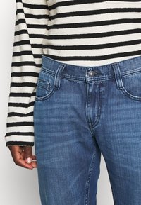 Jeans de mezclilla azul con corte recto y un ligero desgastado. Incluye bolsillos delanteros y cierre de botón. Combinados con una blusa de rayas en blanco y negro.