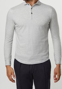 Hombre vistiendo una camiseta polo de manga larga gris claro con tres botones y pantalones azul marino oscuros, de pie contra un fondo liso.