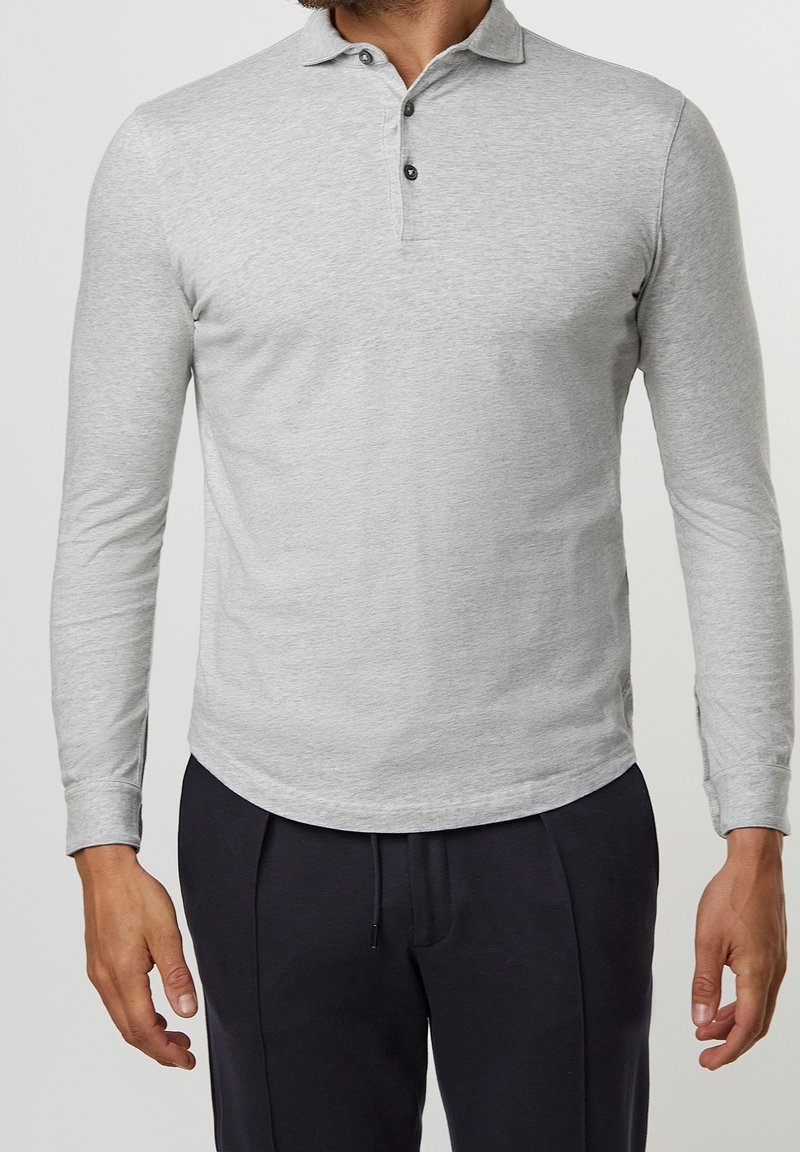 Hombre vistiendo una camiseta polo de manga larga gris claro con tres botones y pantalones azul marino oscuros, de pie contra un fondo liso.