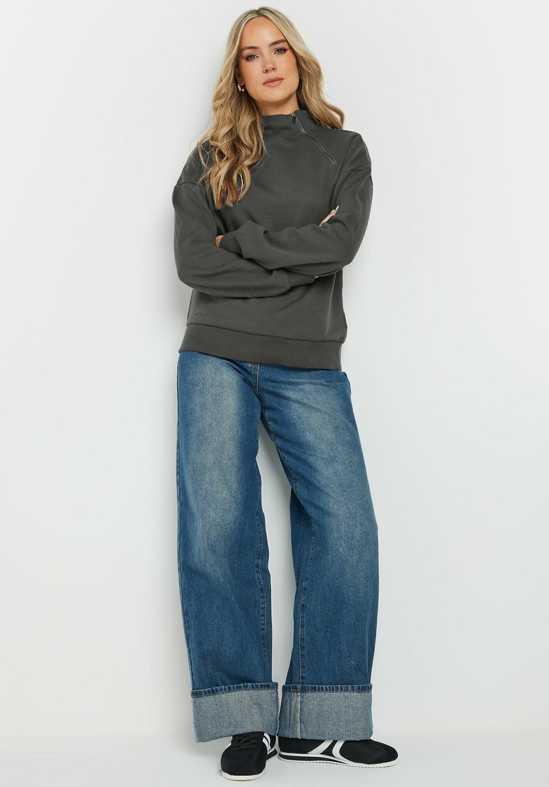 Mörkgrå sweatshirt med halvvippad krage, kombinerad med vida blå denimjeans som har uppvikta muddar och svarta sneakers.