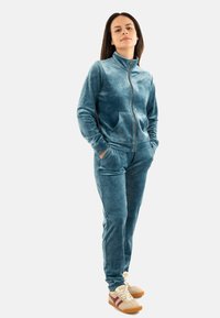 Tuta in velour blu con giacca zip e tasche e pantaloni abbinati. Il tessuto ha un aspetto morbido e strutturato. Scarpe casual incluse.