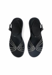 Sandales noires avec un design à sangle croisée, présentant des accents en chaîne argentée. Construites en matériau lisse avec une semelle plate. Sangle réglable.