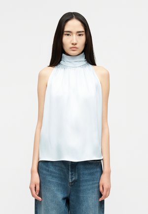 Jeune femme aux longs cheveux noirs, portant un chemisier sans manches bleu clair à col haut et un jean bleu ample, se tenant devant un fond blanc.