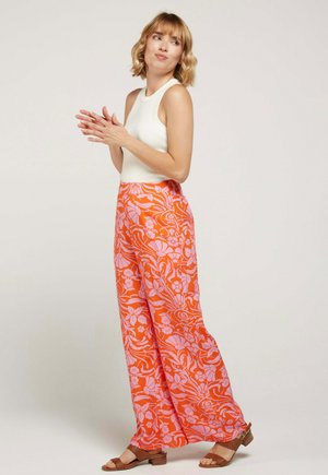 Femme aux cheveux blonds courts portant un haut blanc sans manches, un pantalon orange à jambes larges avec un imprimé floral rose, et des sandales marron, debout, applaudissant.