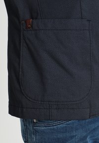 Dunkelblaues Stoffjackett mit strukturierter Oberfläche, ausgestattet mit einer großen aufgenähten Tasche und einem braunen Lederakzent nahe der Taschenöffnung.