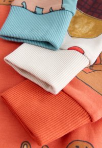 Tre ribbede bomuldærmer i lyseblå, hvid og orange med legende mønstre mod en blød, tekstureret baggrund.