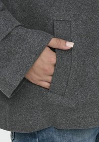 Veste en laine grise à manches larges, avec une main dans une poche latérale. La texture semble douce et légèrement duveteuse.