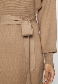 Robe en tricot côtelé beige à manches longues avec une ceinture nouée à la taille, présentée sur la section médiane et le bras droit d'une personne.