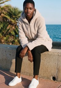 Jaunuolis, vilkintis kreminės spalvos fliiso striukę ir juodus kelnes, sėdi ant betono briaunos prie jūros, fone matyti palmės.