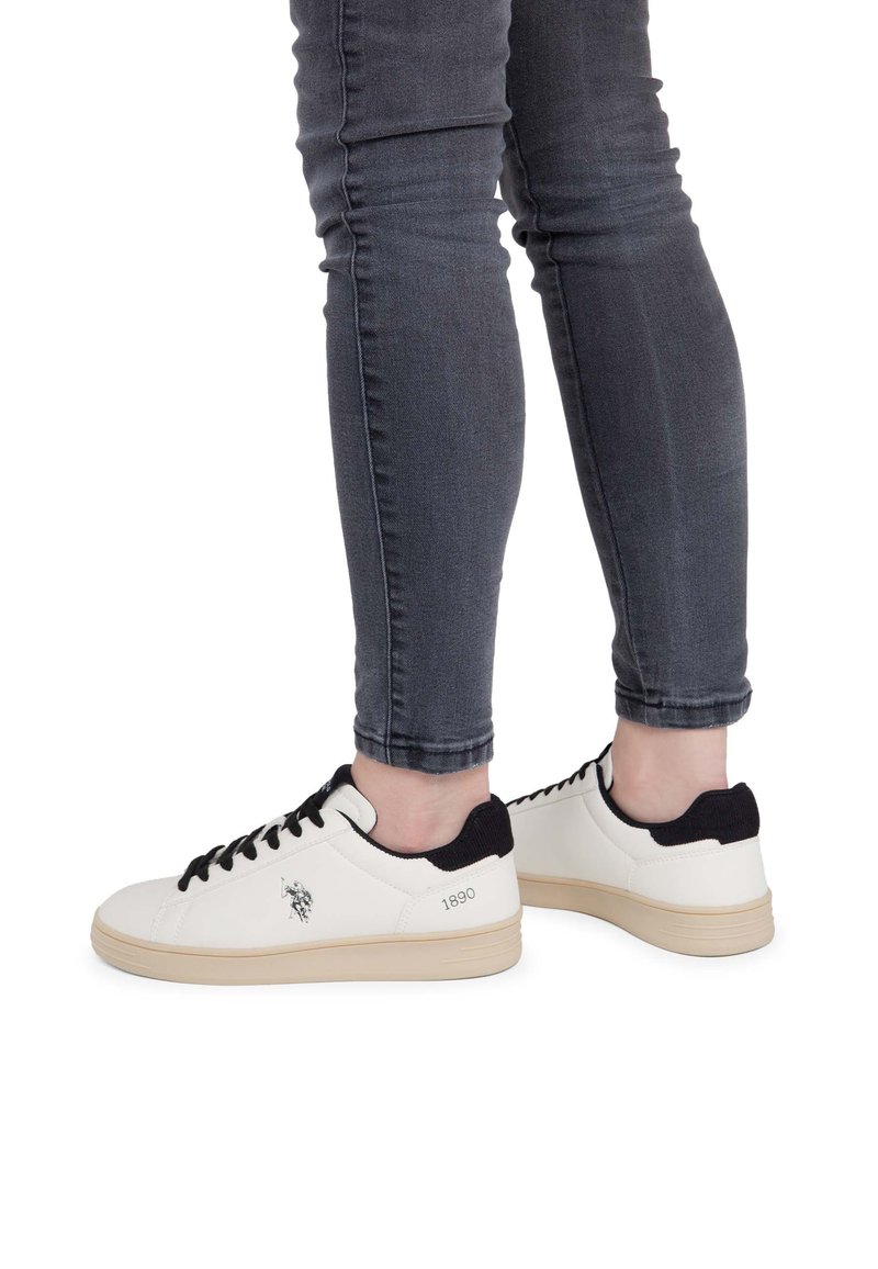 Jambes portant un jean gris foncé moulant et des baskets blanches avec des lacets noirs et un détail noir au talon, debout sur un fond blanc.