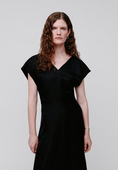 Γυναίκα με καστανά σγουρά μαλλιά φοράει μαύρο φόρεμα με V λαιμόκοψη και ασημένιο βραχιόλι, στέκεται μπροστά σε απλό ανοιχτό γκρι φόντο.