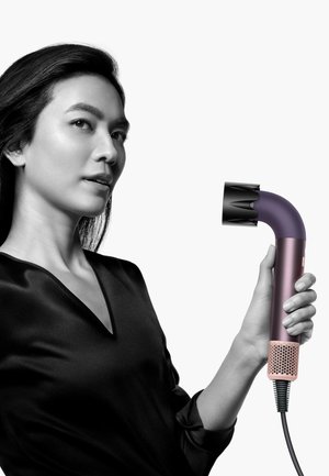 Haartrockner mit geschwungenem Design, ausgestattet mit einem matten lila Griff, einer schwarzen Düse und einer strukturierten rosa Basis. Das Kabel ist schwarz und aufgewickelt.