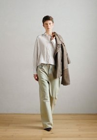 Modelis, ģērbtam gaiši svītrainā īsajā krekla un gaiši zaļajās platajās biksēs, uz pleca tur brūnu ādas jaku.