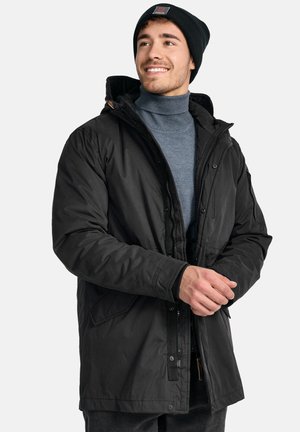 Manteau noir rembourré avec une capuche, fermeture éclair à l'avant et boutons pression. Doté de poches latérales et d'un col texturé. Porté par-dessus un pull à col roulé bleu.