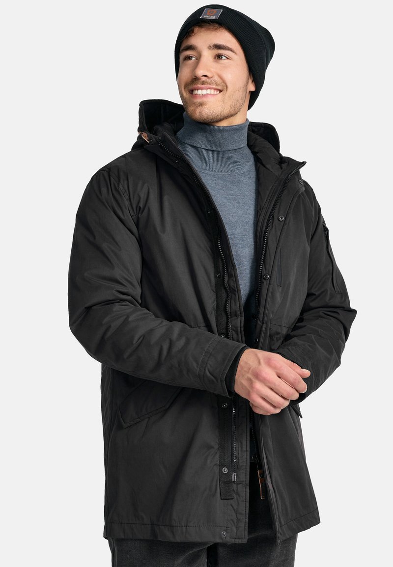 Abrigo negro acolchado con capucha, cremallera frontal y botones de broche. Presenta bolsillos laterales y un cuello texturizado. Llevado sobre un suéter de cuello alto azul.