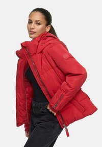 Red puffer jacket with a high collar, zippered front, and side pockets. Features quilted texture and logo on the sleeve. Worn over a black top.