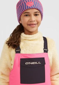 Roze en zwarte skibroek met een zak, gecombineerd met een crèmekleurige fuzzy jas. Een meerkleurige muts met een logo.