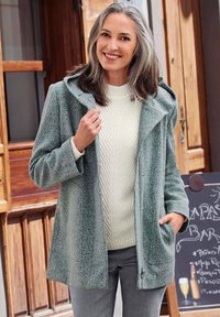 Hellblauer Mantel mit Kapuze, aus strukturiertem Stoff, mit vorderem Taschen und Reißverschluss, getragen über einem cremefarbenen, kabelgestrickten Pullover.