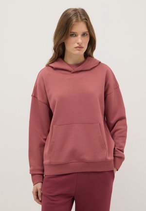 Joven con cabello castaño claro que lleva una sudadera con capucha color rosa polvo y pantalones a juego, de pie contra un fondo claro y liso.