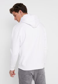 Hvit hettegenser med hette, ribbestrikkede mansjetter og kant, samt myk, teksturert stoff. Bært med grå jeans, som viser en uformell passform.
