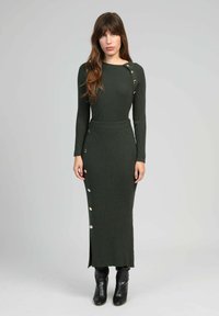 Robe en tricot côtelé de couleur vert foncé, avec des manches longues, des boutons sur le côté, une taille cintrée et une fente à l'avant, assortie à des bottines noires.