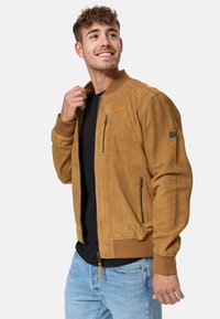 Tan suede bomber jacket with a ribbed collar, zippered pockets, and textured finish. Paired with a black shirt and light blue jeans.