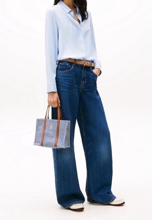 Vrouw draagt lichtblauwe blouse en donkerblauwe wijde jeans, houdt een kleine blauw-wit gestreepte draagtas met bruine handvatten vast.