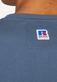 Sininen puuvillapaita, jossa on ribbetty kaula-aukko. Takana on valkoinen yksityiskohta, jossa on monivärinen "R" ja "BOSS" -logo.