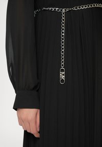 Κοντινό πλάνο ενός χεριού που φορά ένα μαύρο πλισέ φόρεμα με διαφανείς μακρύ μανίκια και μια ασημένια αλυσίδα μέσης με κρεμαστό ορθογώνιο γούρι.