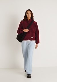 Μπουφάν από φλις μπορντό με φερμουάρ, δύο μπροστινές τσέπες, συνδυασμένο με ανοιχτό μπλε τζιν παντελόνι φαρδιάς γραμμής και μαύρα παπούτσια, κρατώντας μια μαύρη τσάντα.