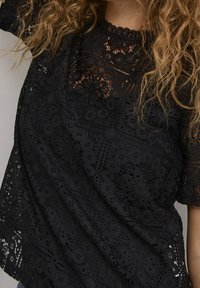 Svart spetsblus med intrikata blommönster, vågig halsringning och korta ärmar. Texturerad design med en lös passform.