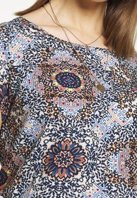 Túnica de estampado floral en crema, con intrincados diseños en marino, azul y naranja. Cuello redondo con mangas cortas, tejido ligero.