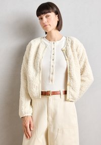 Romantische crèmekleurige nepbontjas met een ronde kraag, voor knopen en geribbelde zoom, gedragen over een witte geribbelde top en lichtbeige broek. Inclusief een bruine riem.
